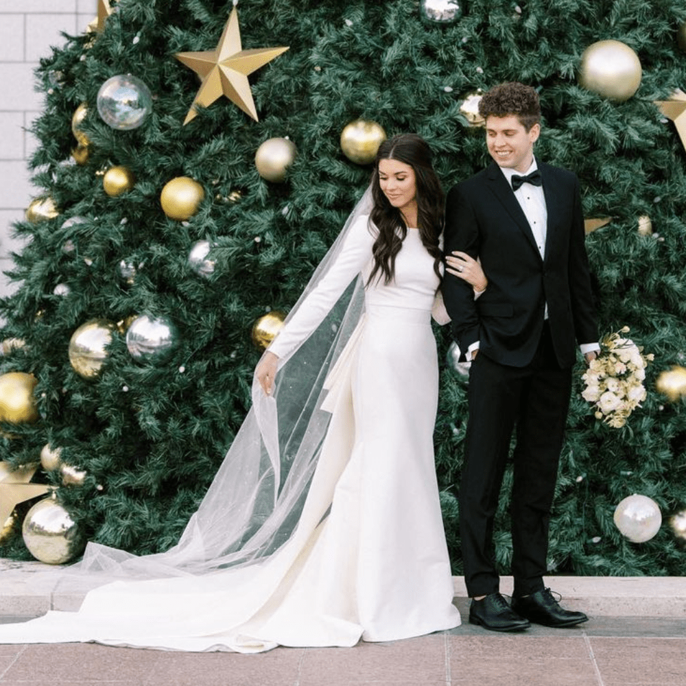 Couple posing in front of a Christmas tree; woman in wedding dress, man in tuxedo.