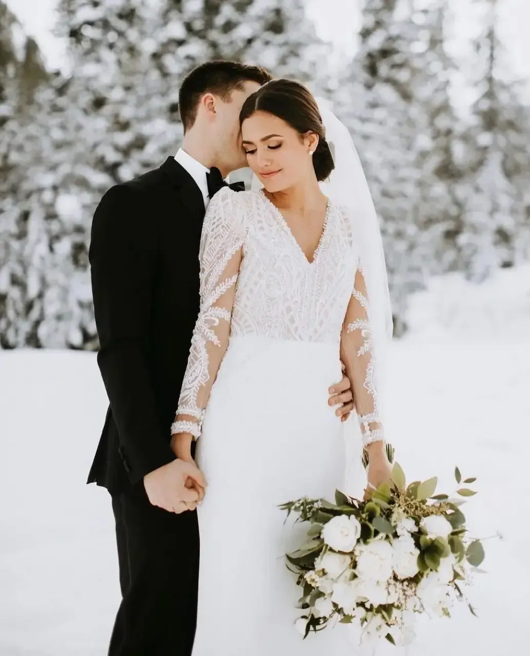 Couple embracing in snow; bride in white dress with bouquet, groom in black suit.