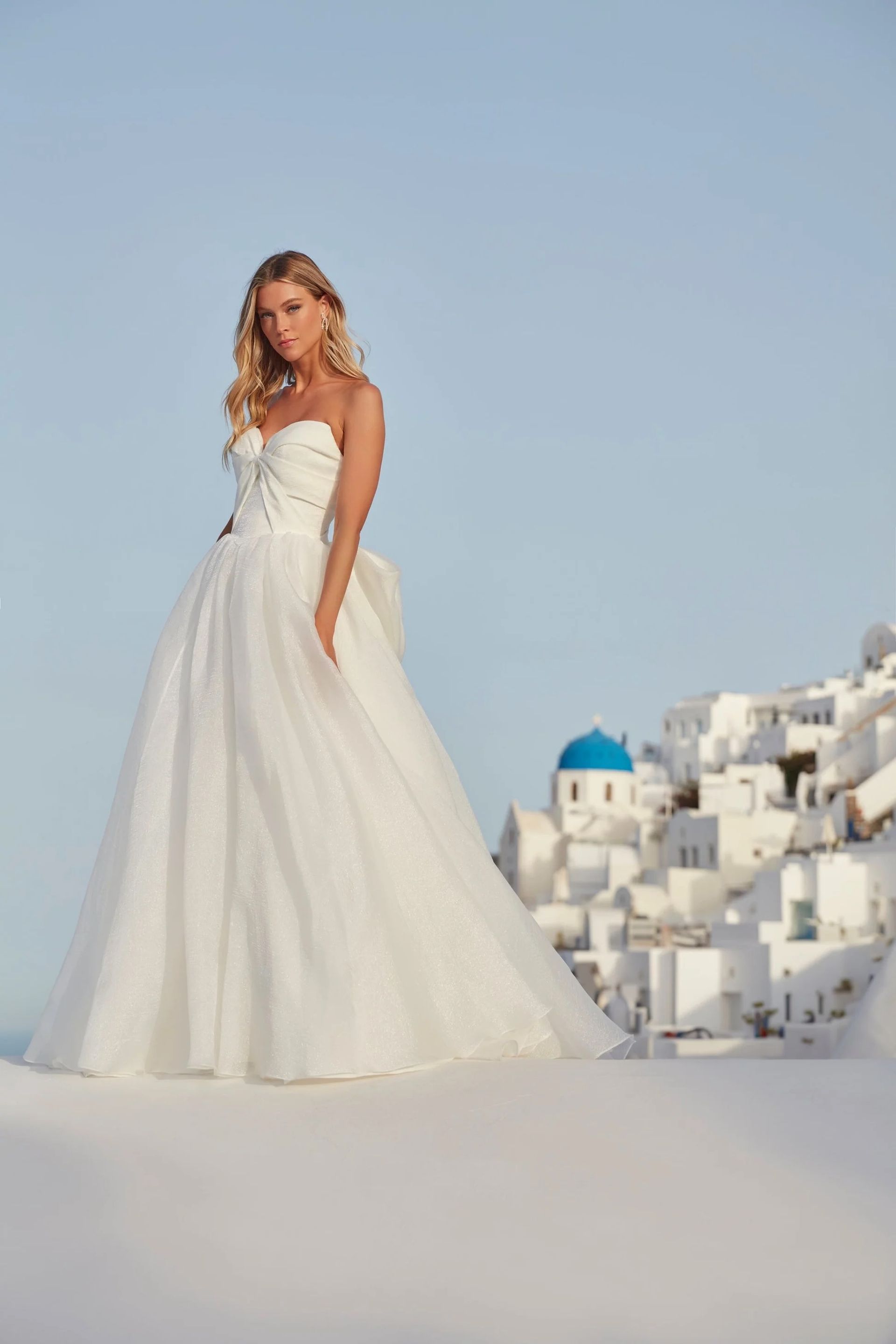 Woman in a white wedding dress posing on a white surface with a background of white buildings and a blue sky.