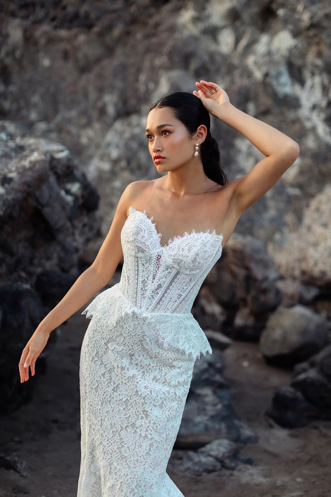 Woman in white lace strapless gown poses outdoors with hand in hair, looking away.