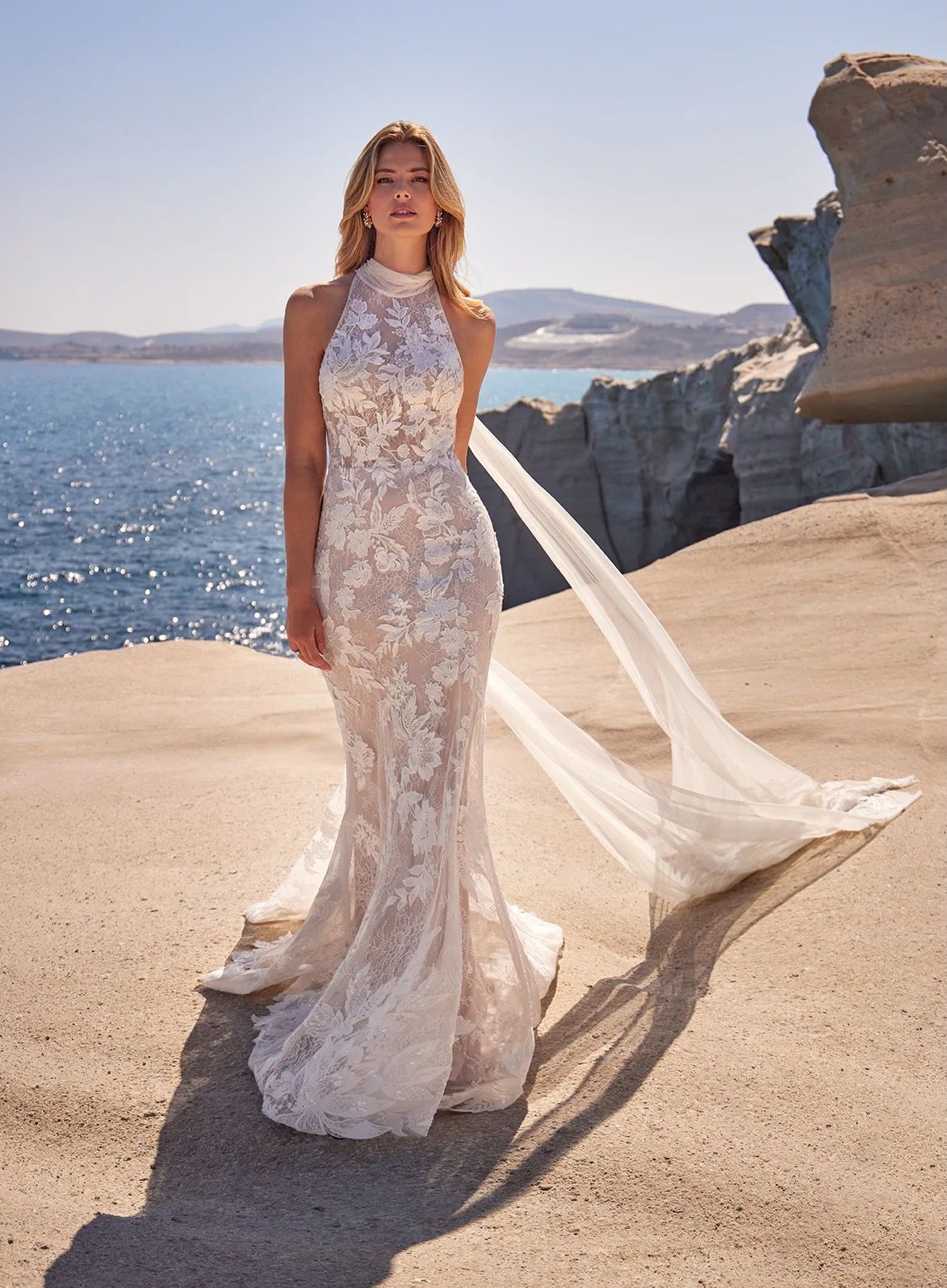 Woman in a white lace wedding dress with halter neckline, standing on a sandy cliff overlooking the sea.