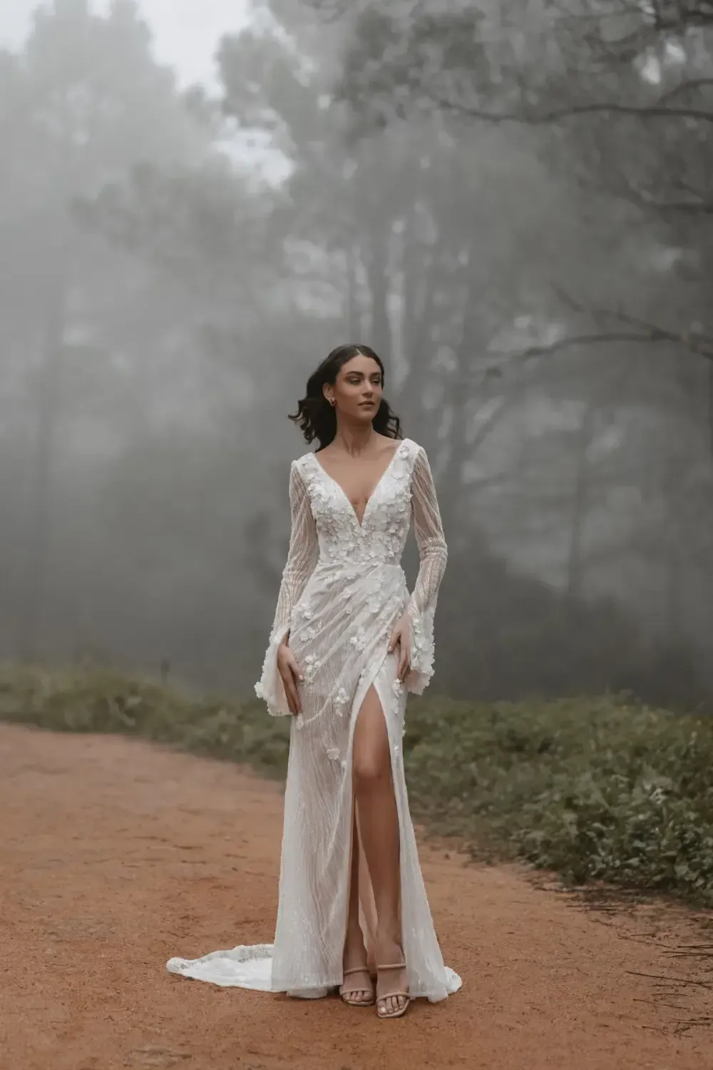 Woman in a white wedding dress stands on a dirt path in a foggy forest.