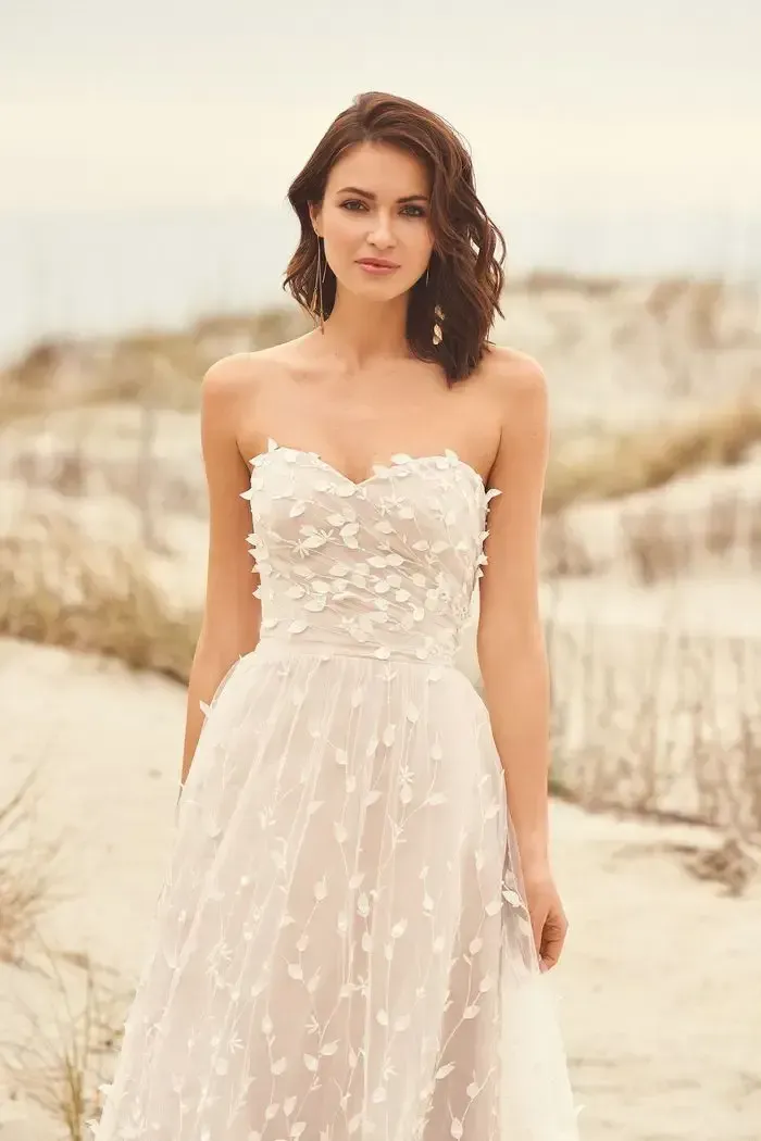 Woman in strapless white wedding dress with floral embellishments on a sandy beach.