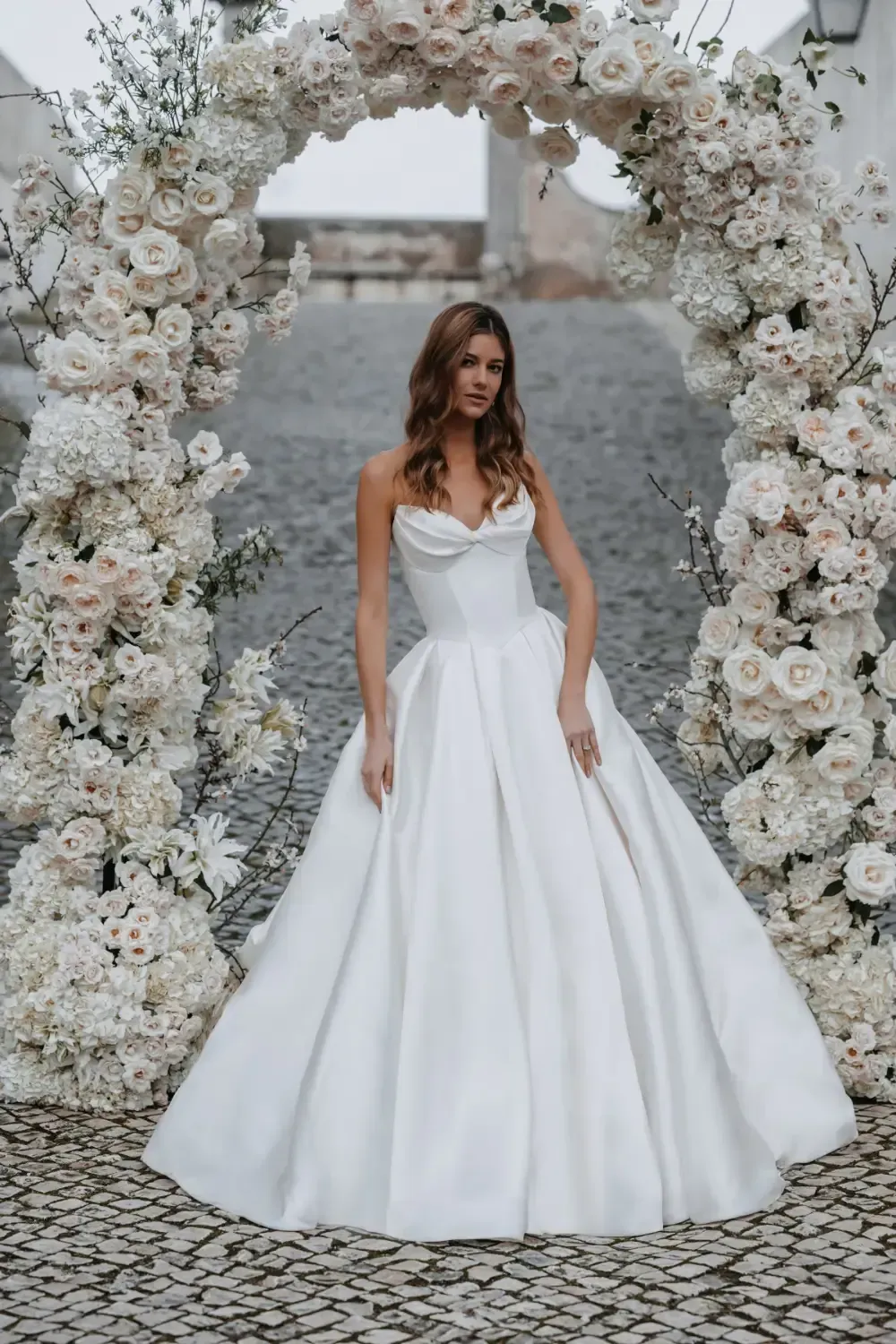 Woman in white wedding dress stands before floral arch.