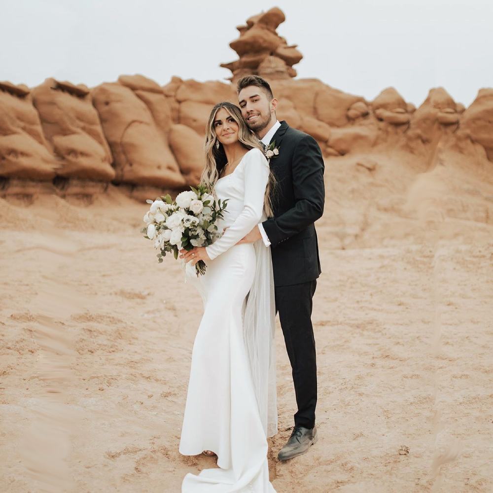 Couple poses for wedding photo in a desert landscape; bride in white dress, groom in black suit.