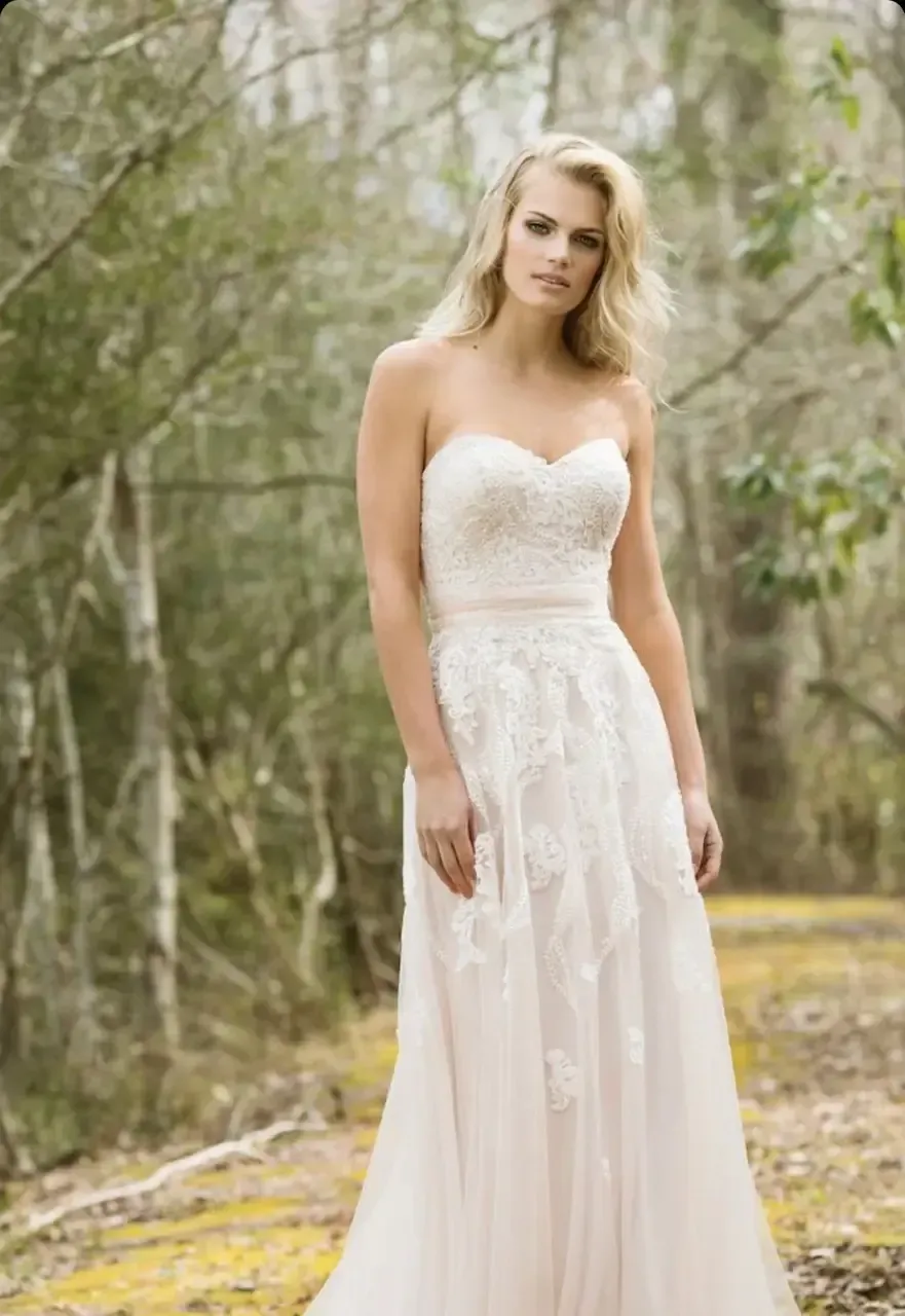 Woman in strapless wedding dress, standing outdoors in front of trees.