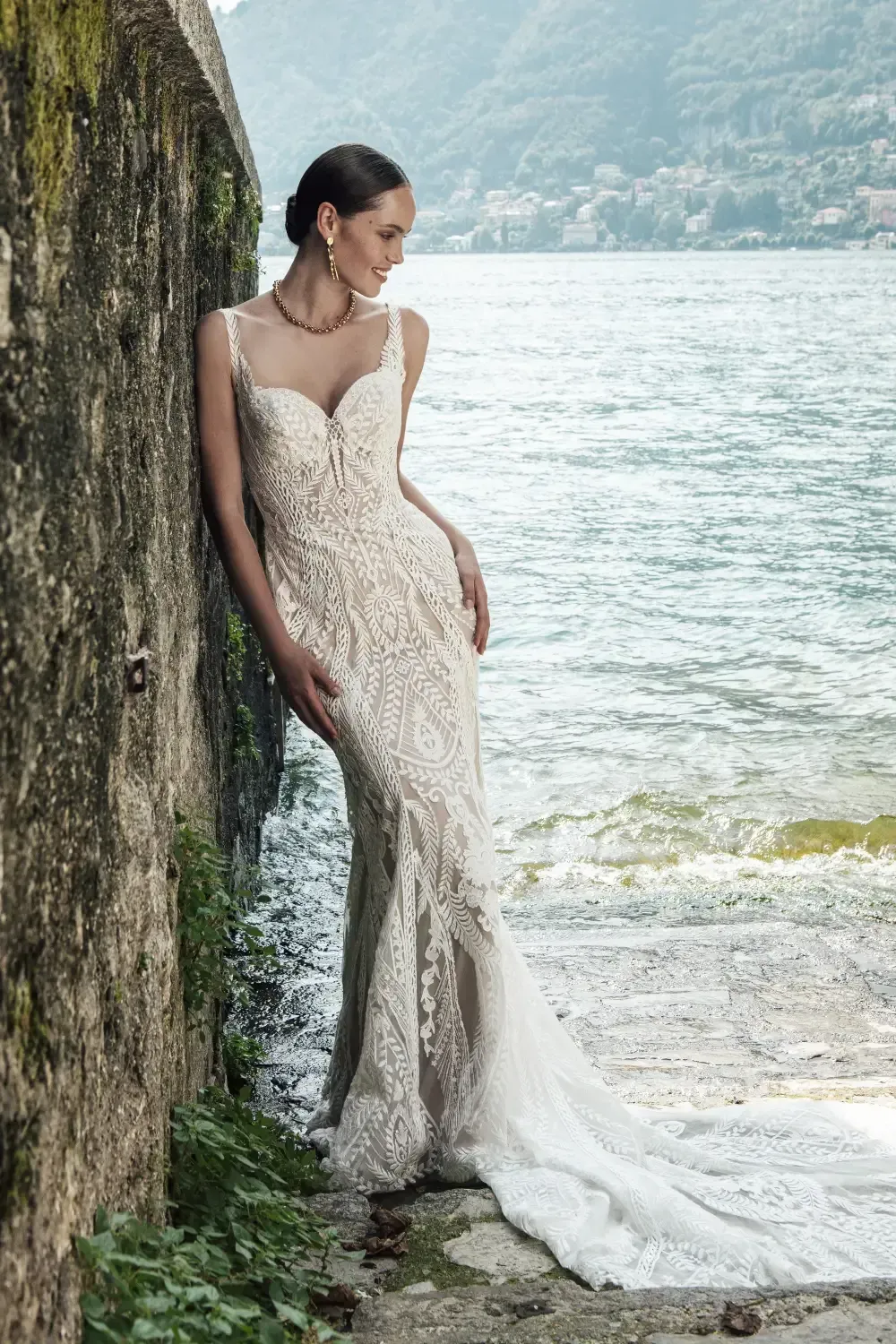 Woman in lace wedding dress leans on a stone wall by a lake, looking off-camera.