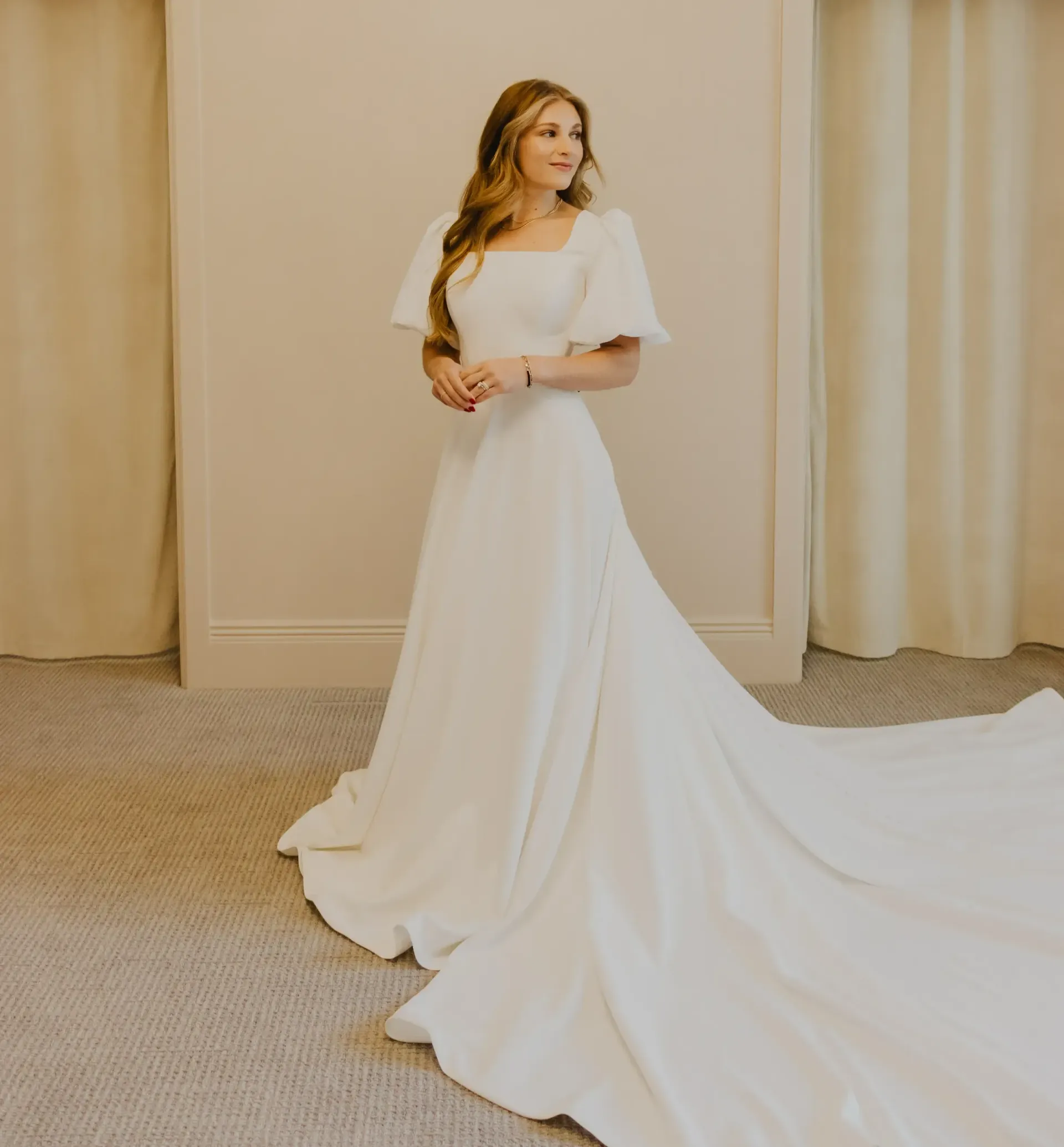 Woman in white wedding dress with long train, standing near curtains, looking away.