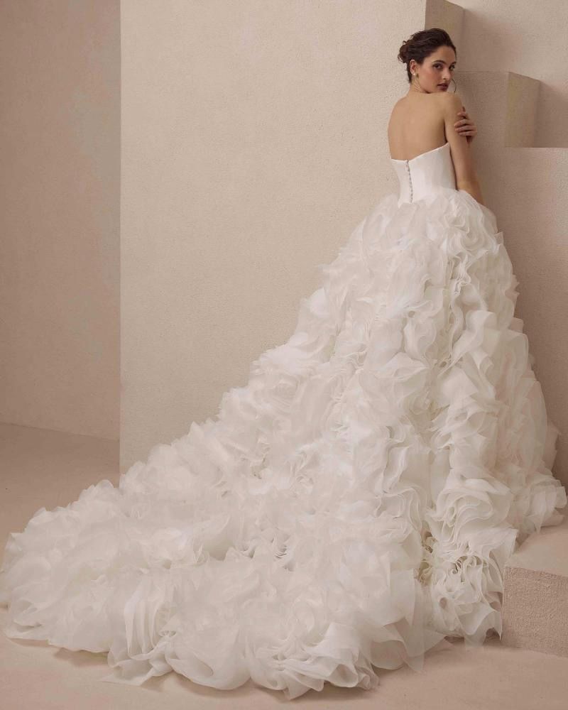 Woman in a strapless white wedding gown with a ruffled skirt and long train, standing against a wall.