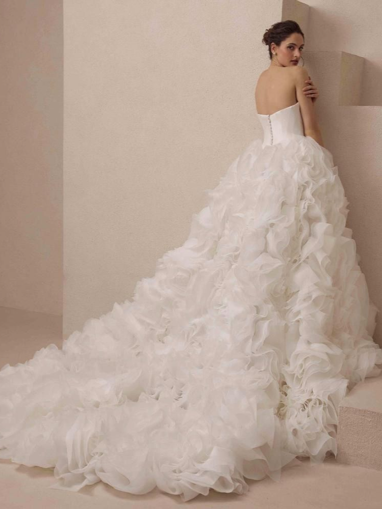 Woman in a strapless white wedding gown with a ruffled skirt and long train, standing against a wall.