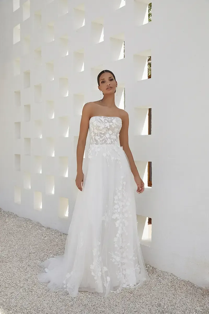 Woman in strapless wedding dress, standing against a white wall with rectangular cutouts.