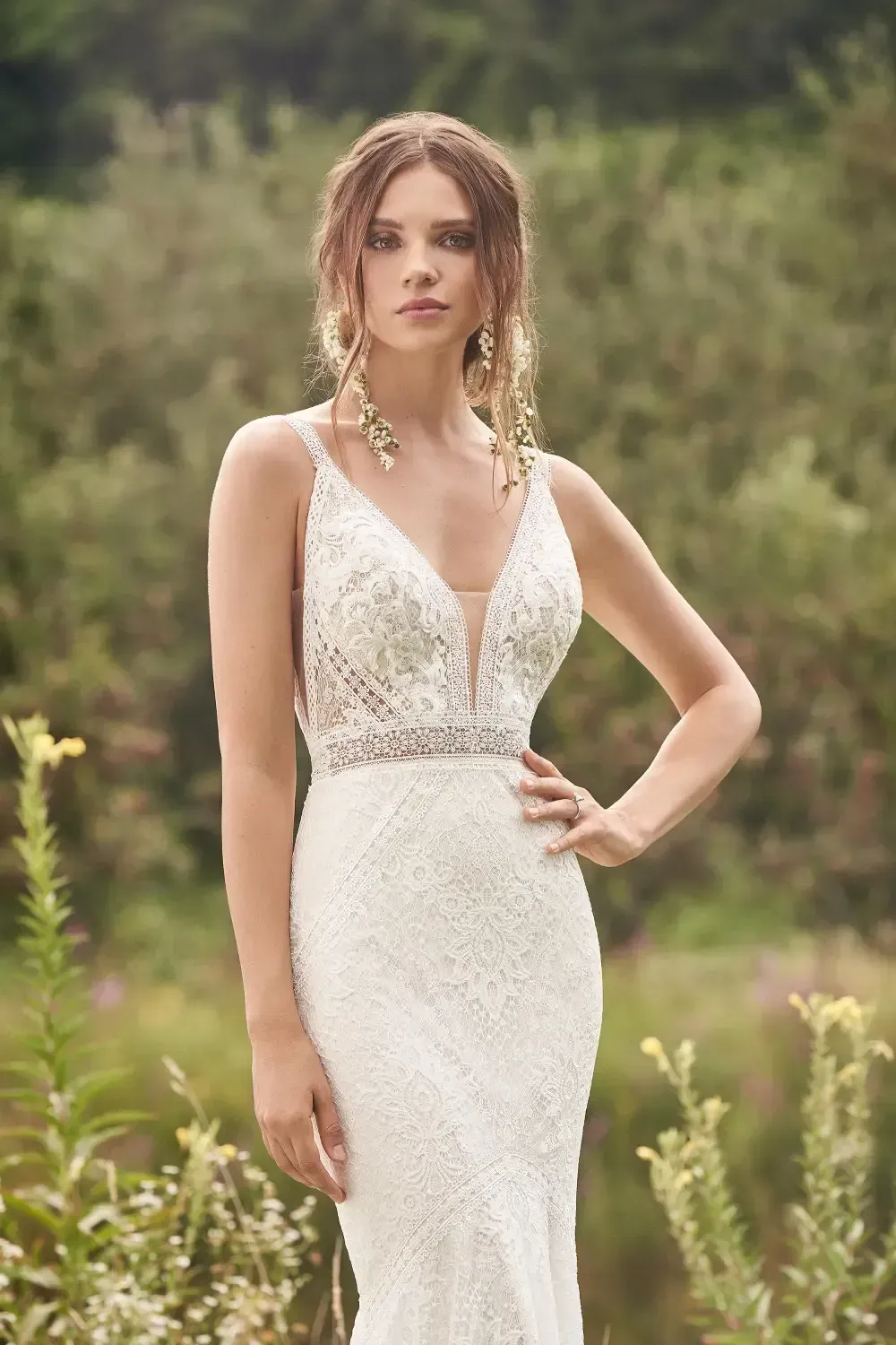 Woman in white, beaded wedding dress with plunging neckline, hand on hip, outdoor setting.