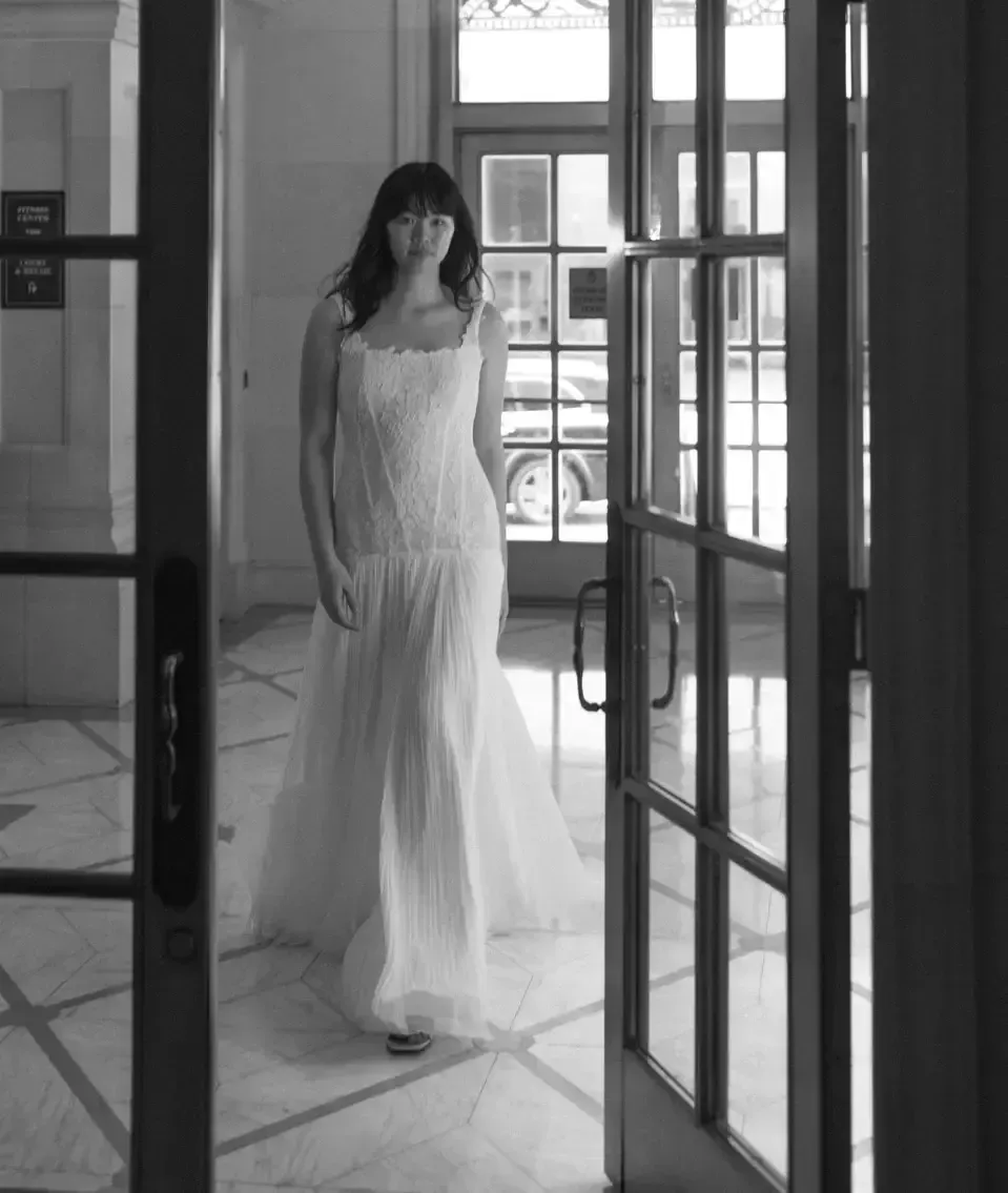 Woman in a white wedding dress walks through a doorway with glass panels and a view of a street.