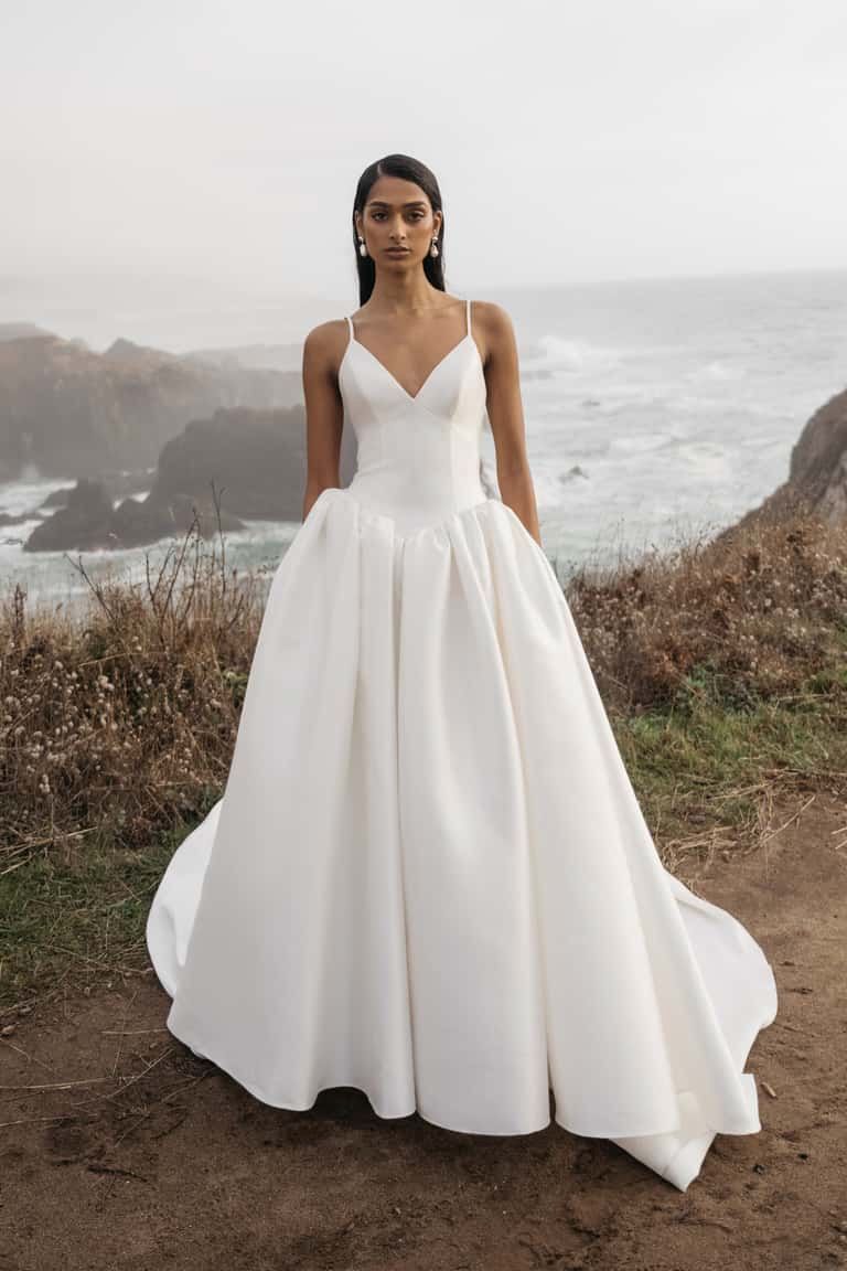 Woman in white satin wedding dress standing near ocean cliffs.