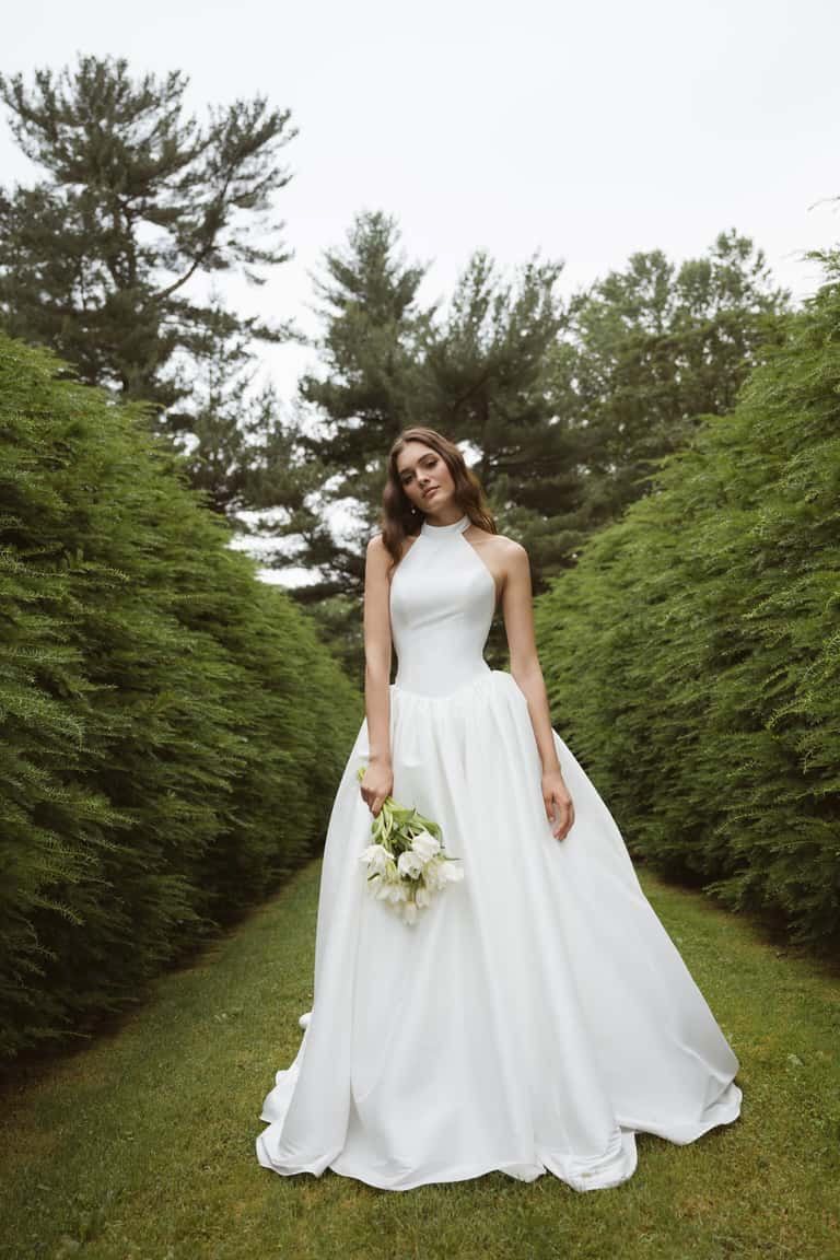 Woman in white wedding dress holding flowers, standing on grassy path lined with hedges.