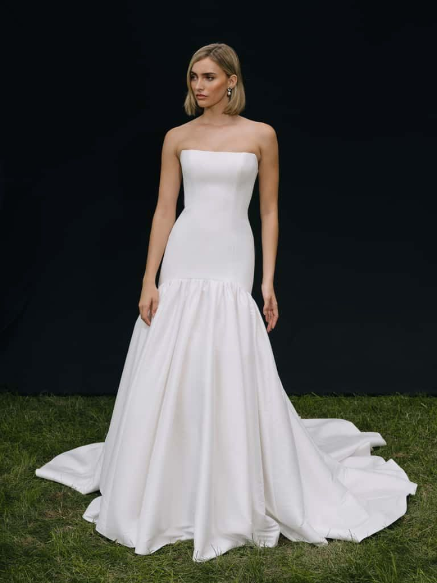 Woman wearing a white strapless mermaid-style wedding gown, standing on grass. Black background.