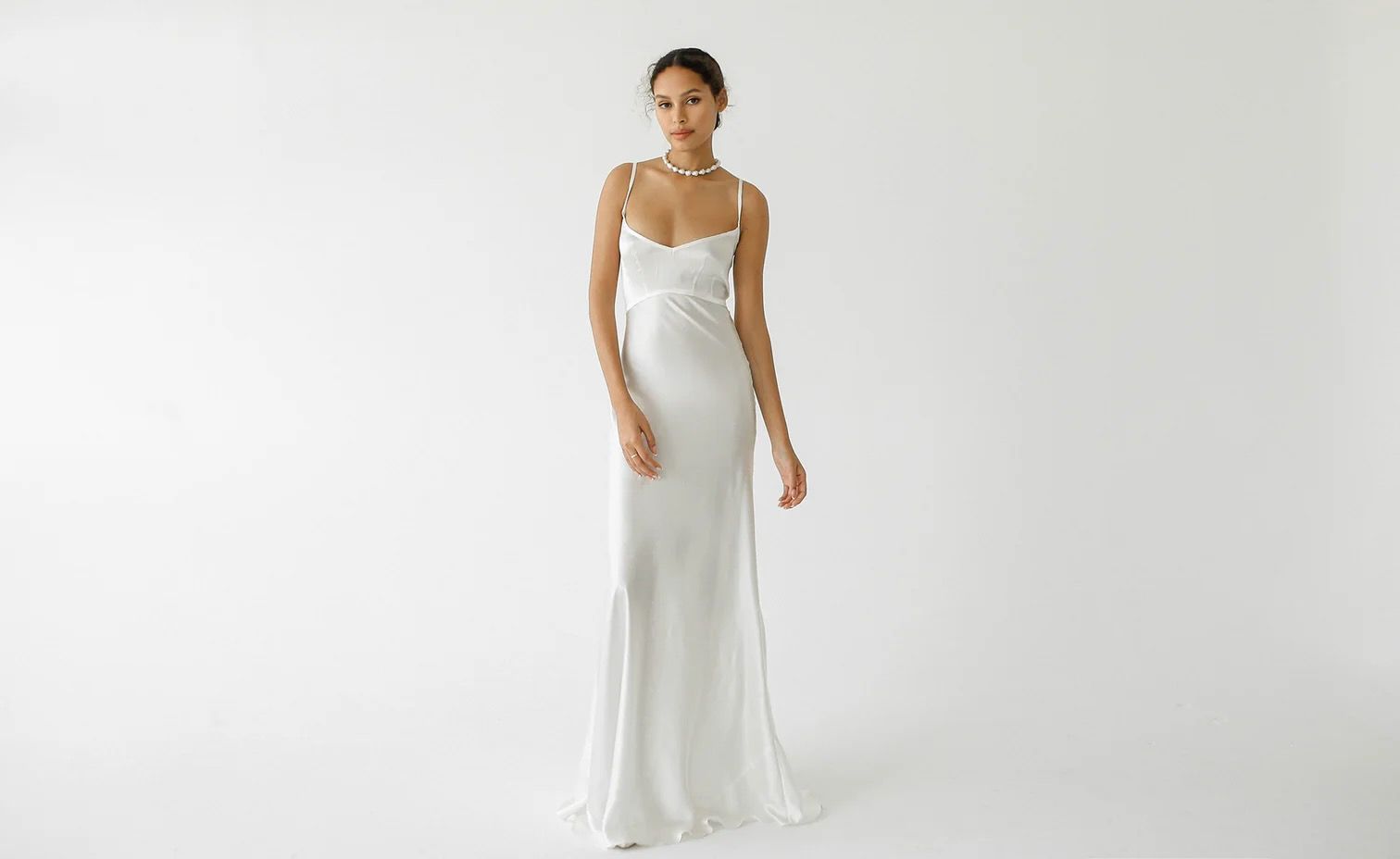 Woman in a white slip dress poses against a white background; wearing a pearl necklace.
