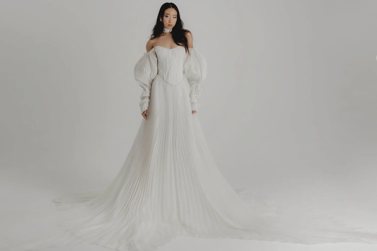 Woman in white off-the-shoulder wedding gown with long train, posing in a studio.