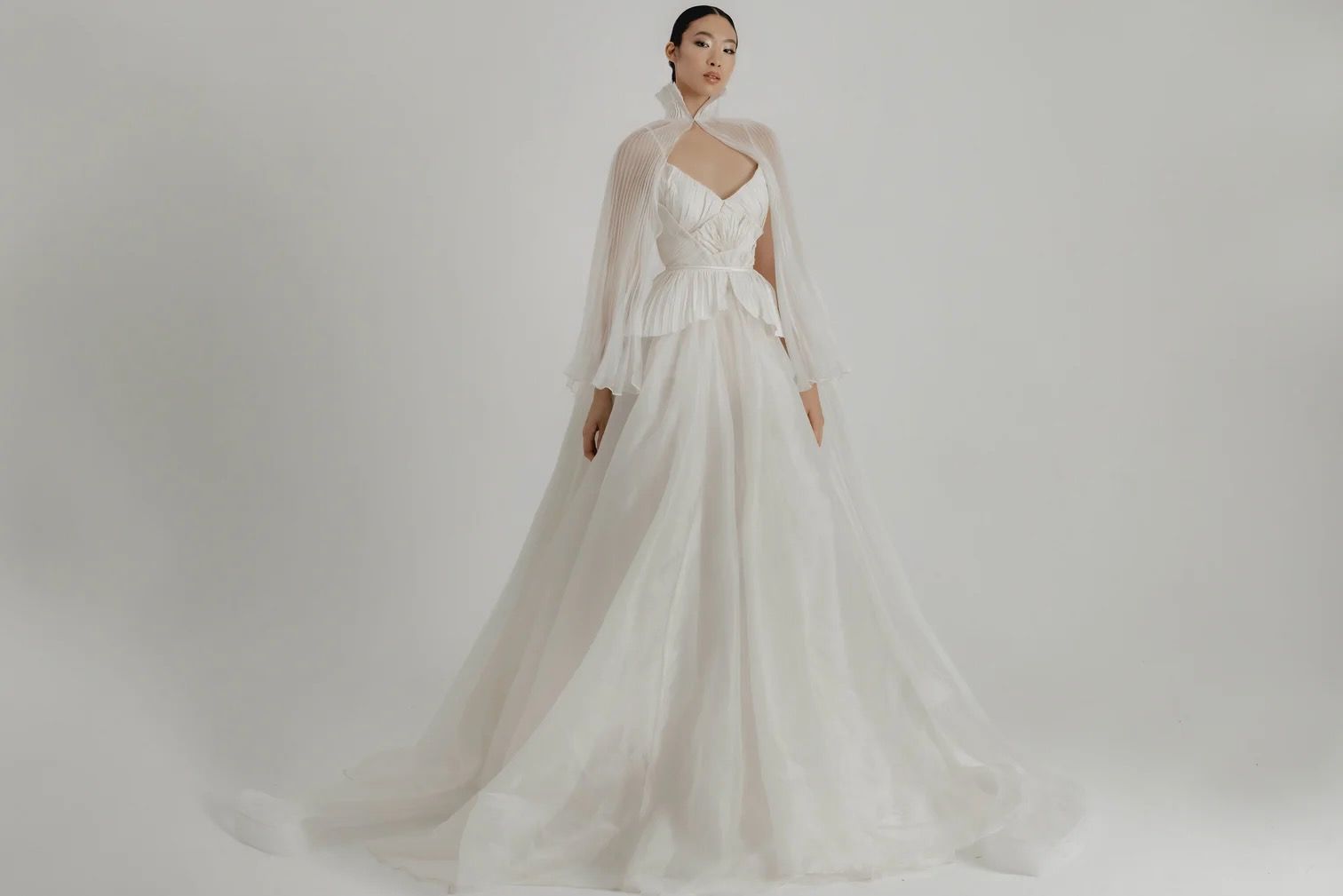 Woman in a white wedding gown with a cape, posing in a studio.