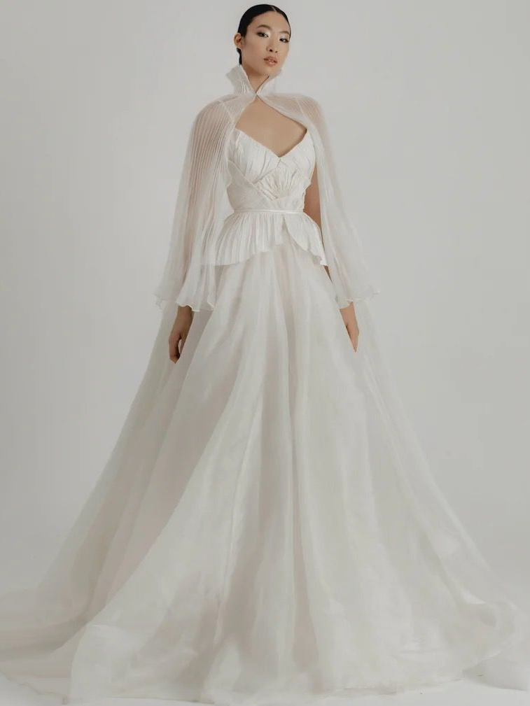 Woman in a white wedding gown with a cape, posing in a studio.