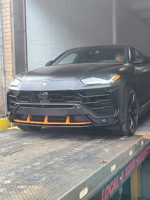 Dark gray Lamborghini Urus SUV on a tow truck, with orange trim, parked indoors.
