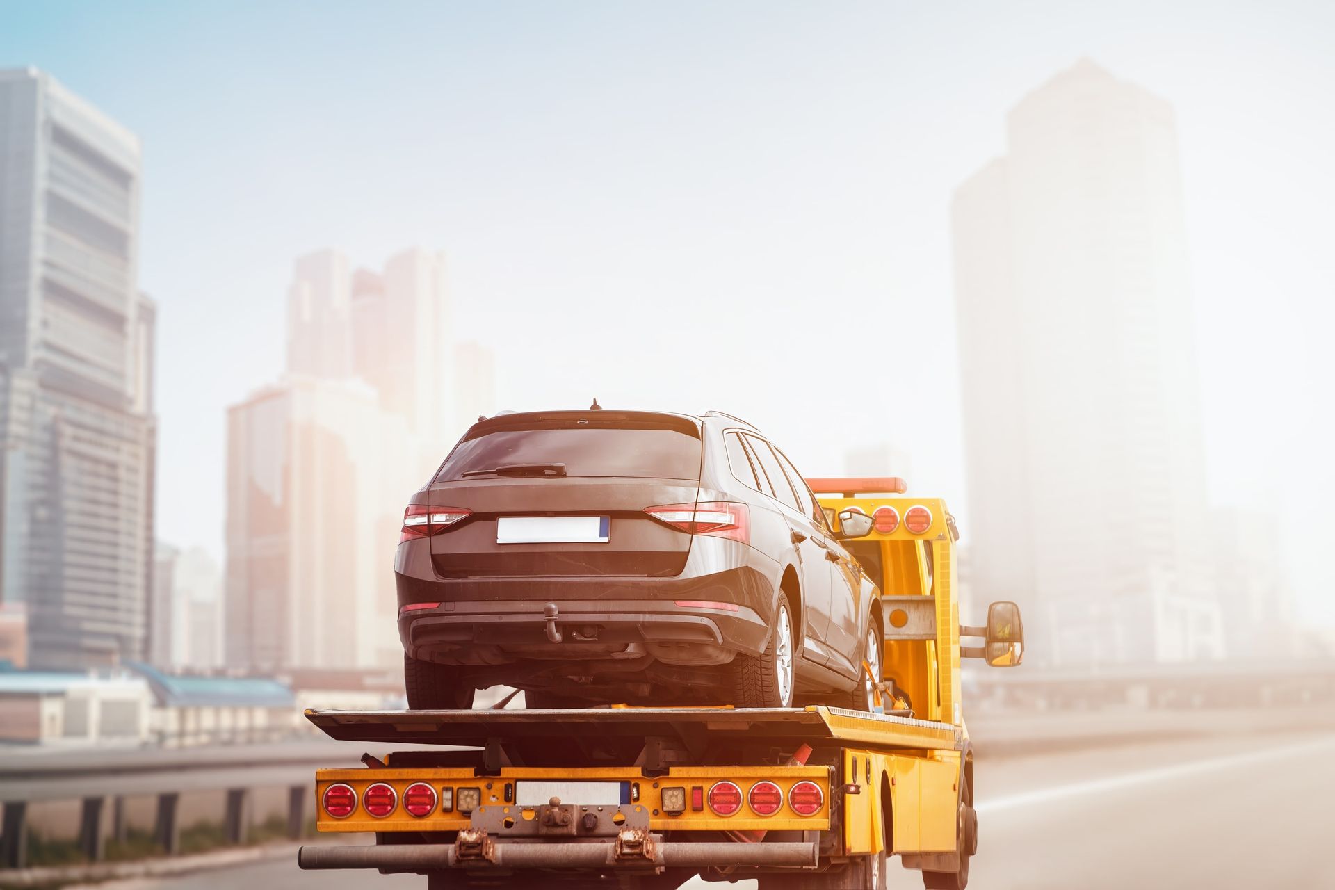 Black SUV being towed by a yellow tow truck on a highway with city buildings in the background.