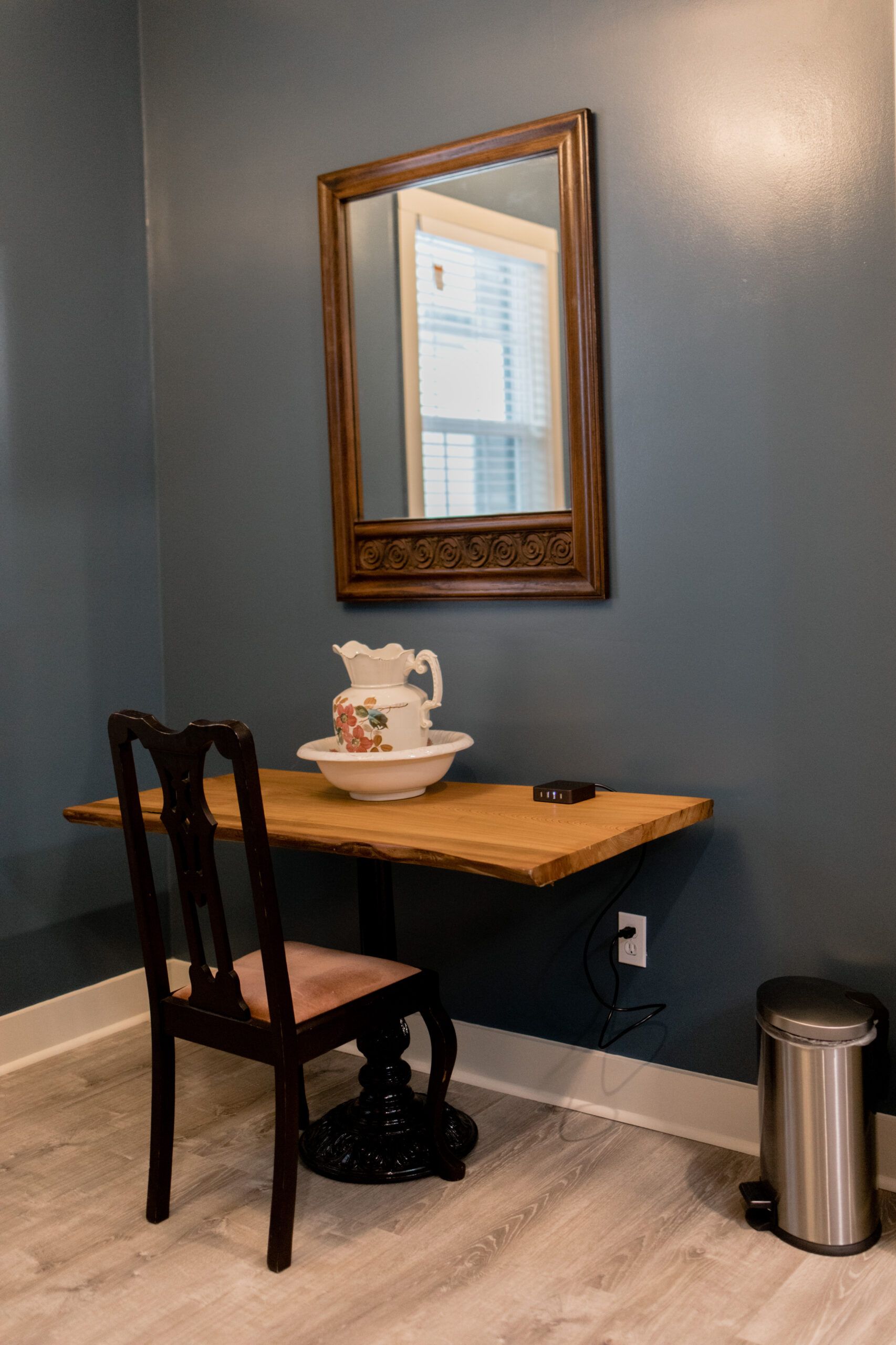 A room with a table , chair , mirror and trash can.