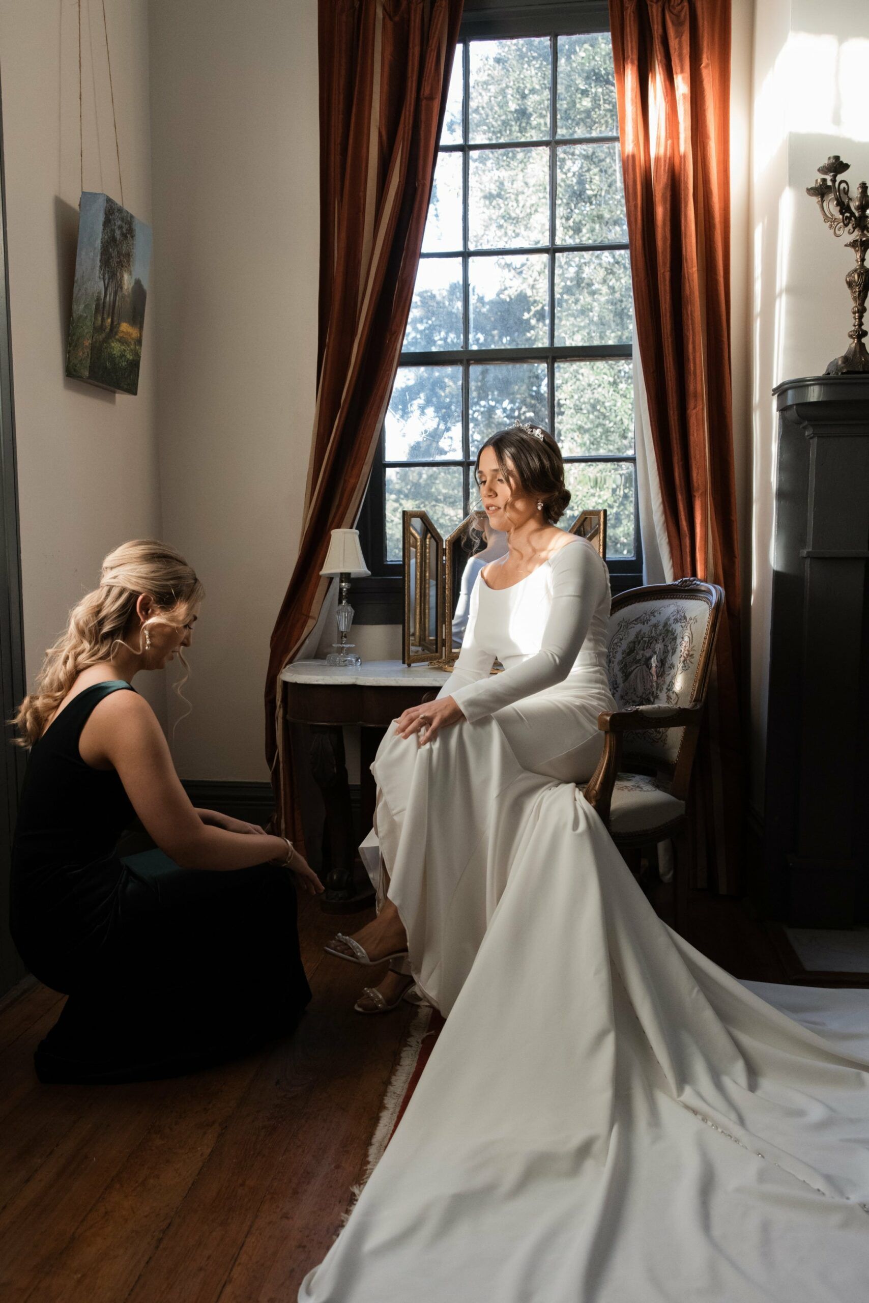 A woman in a wedding dress is sitting on a chair in front of a window.
