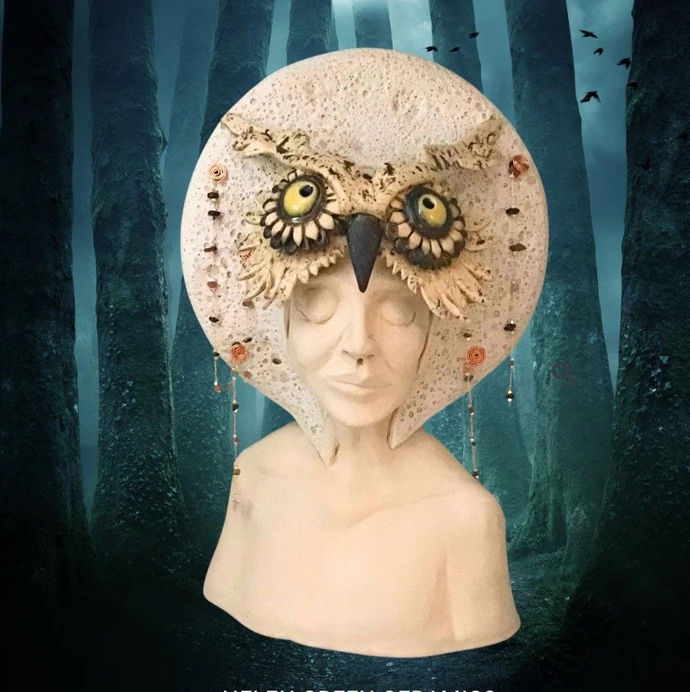 Photo of a ceramic head and shoulders; a mystical being, young female human wearing a headdress with an owl head at the centre, adorned with gold chains with gemstones; the background is a dark forest.