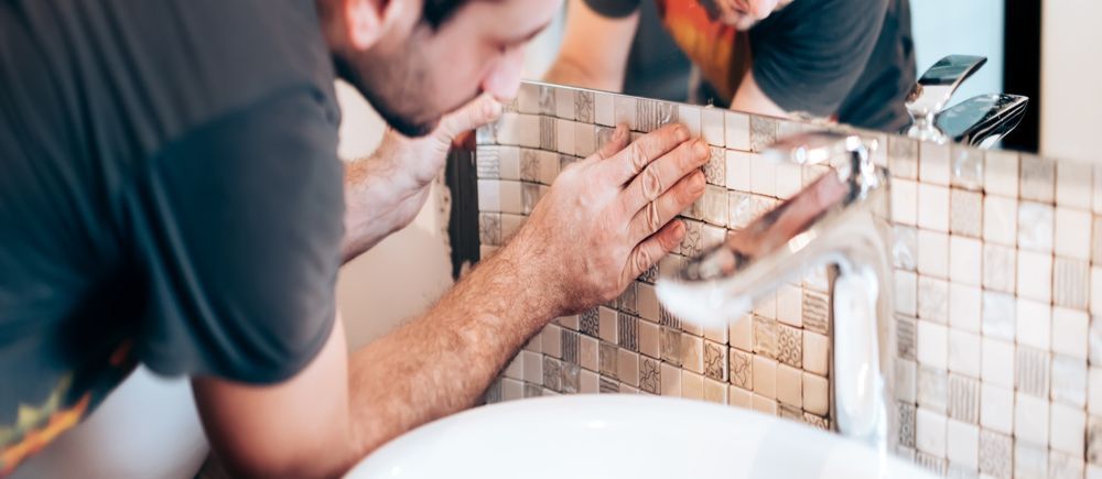 A person installs mosaic tiles near a faucet and sink.