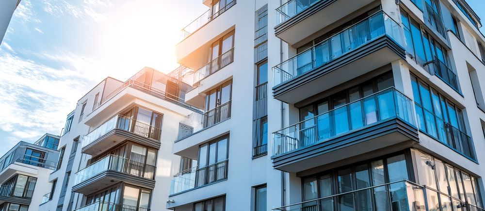 Modern white apartment building with balconies, bright sun, and blue sky.