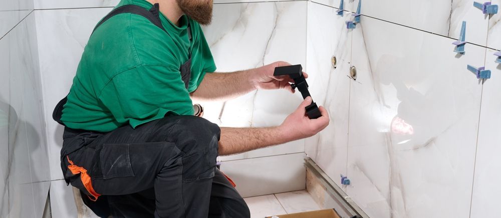 A person kneels, installing tiles in a bathroom. They hold a black tool. The walls are white.