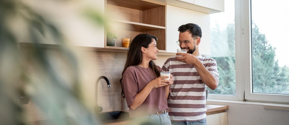 Couple in kitchen, drinking from glasses, looking at each other, smiling, sunlight through window.