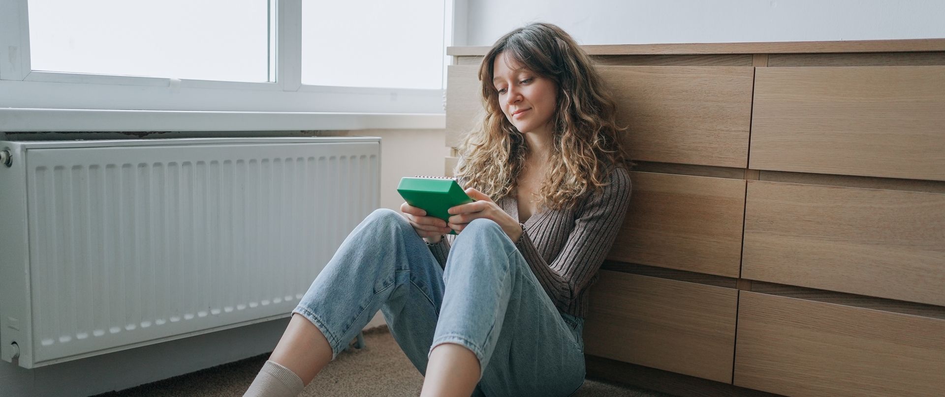 Woman sitting on the floor by a wooden dresser, using a green calculator.  She is wearing jeans and a sweater.