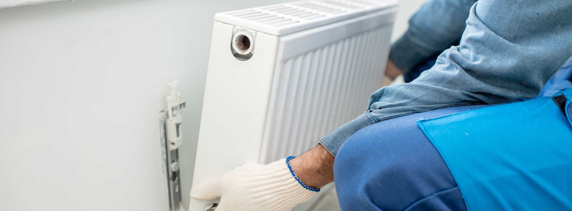 A person in blue overalls installing a white radiator on a white wall.