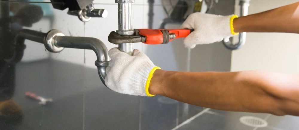 A plumber in gloves uses a wrench to tighten a pipe beneath a sink.