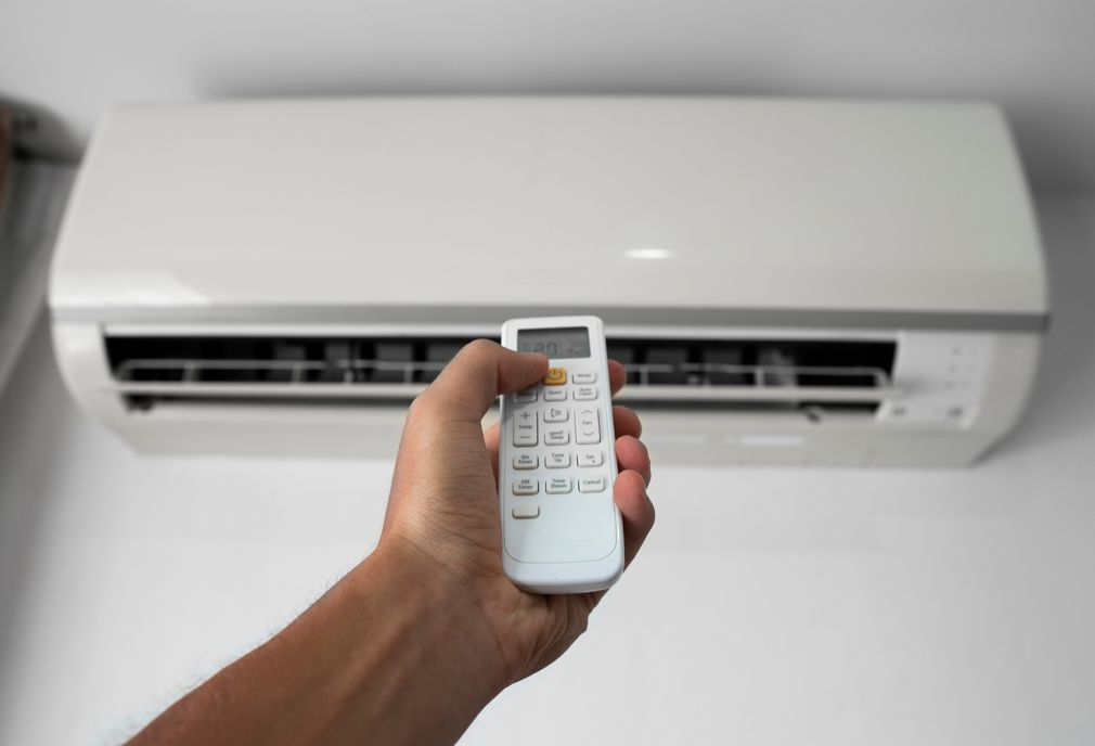 Hand holding AC remote pointing towards a mounted white air conditioner.