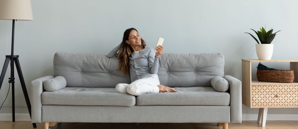 Woman on a couch looks at her phone, smiling. A side table with a plant and a lamp are nearby.