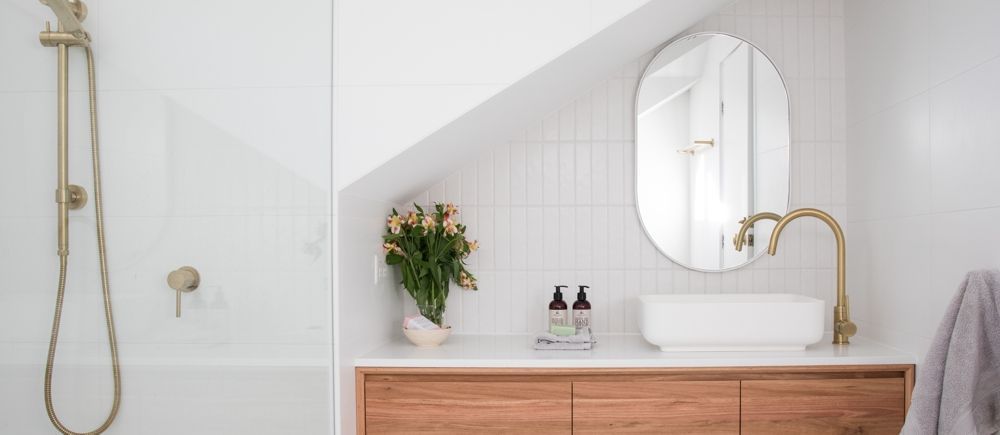Bathroom with wooden vanity, gold faucet, oval mirror, white sink, and flowers.