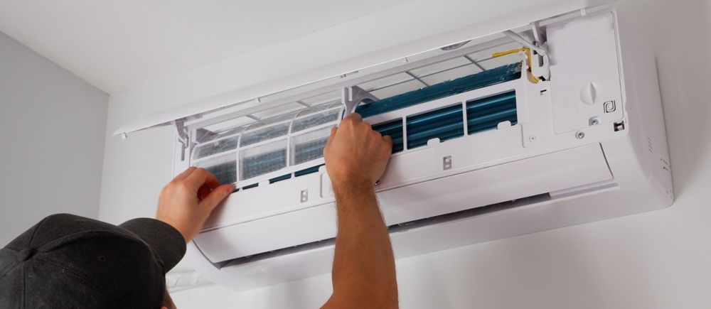 Person cleaning air conditioner filter. Hands near the white unit, indoors.