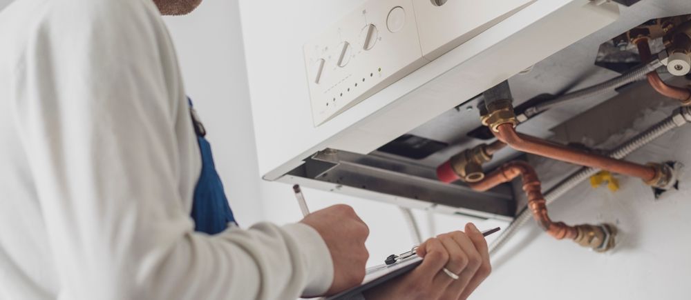 A person in a white shirt and blue overalls checks a boiler, taking notes on a clipboard.