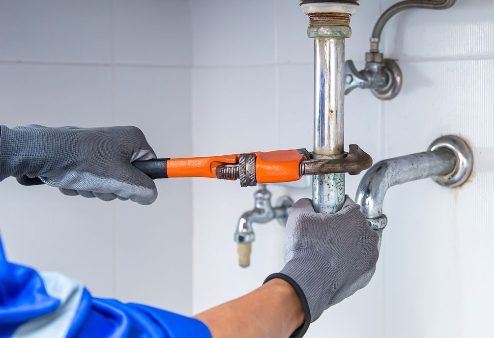 Plumber in gloves using wrench to repair sink pipes in a white tiled bathroom.