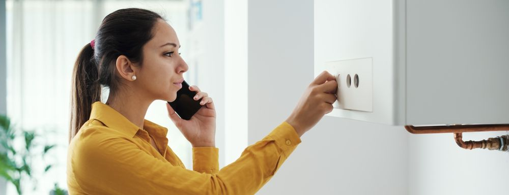 Woman on phone adjusting a white control panel on a boiler. Yellow shirt in a white room.