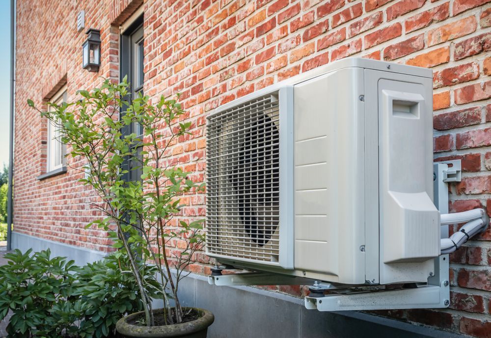 White heat pump unit mounted on a brick exterior wall, with nearby potted plant.
