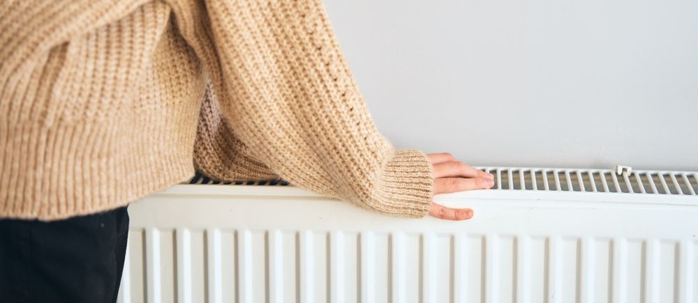 A person wearing a tan sweater touches a white radiator, likely to check its warmth.