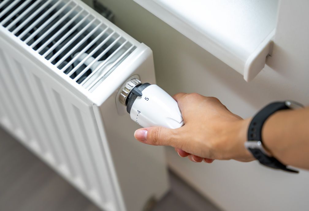 Person's hand turning a white radiator valve. The radiator is white and mounted on a wall near a window.