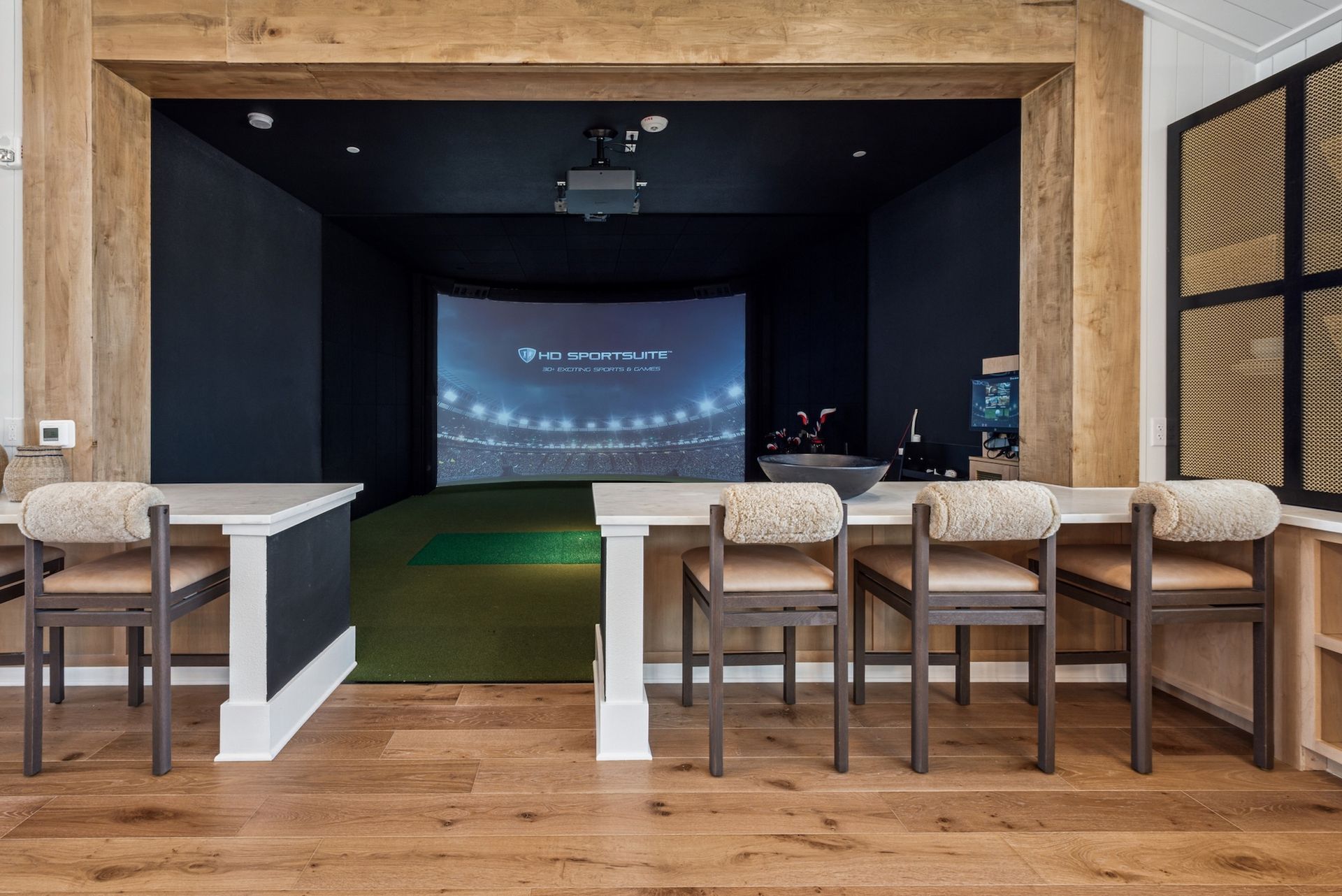 Golf simulator room with screen, green turf, tables, and chairs.