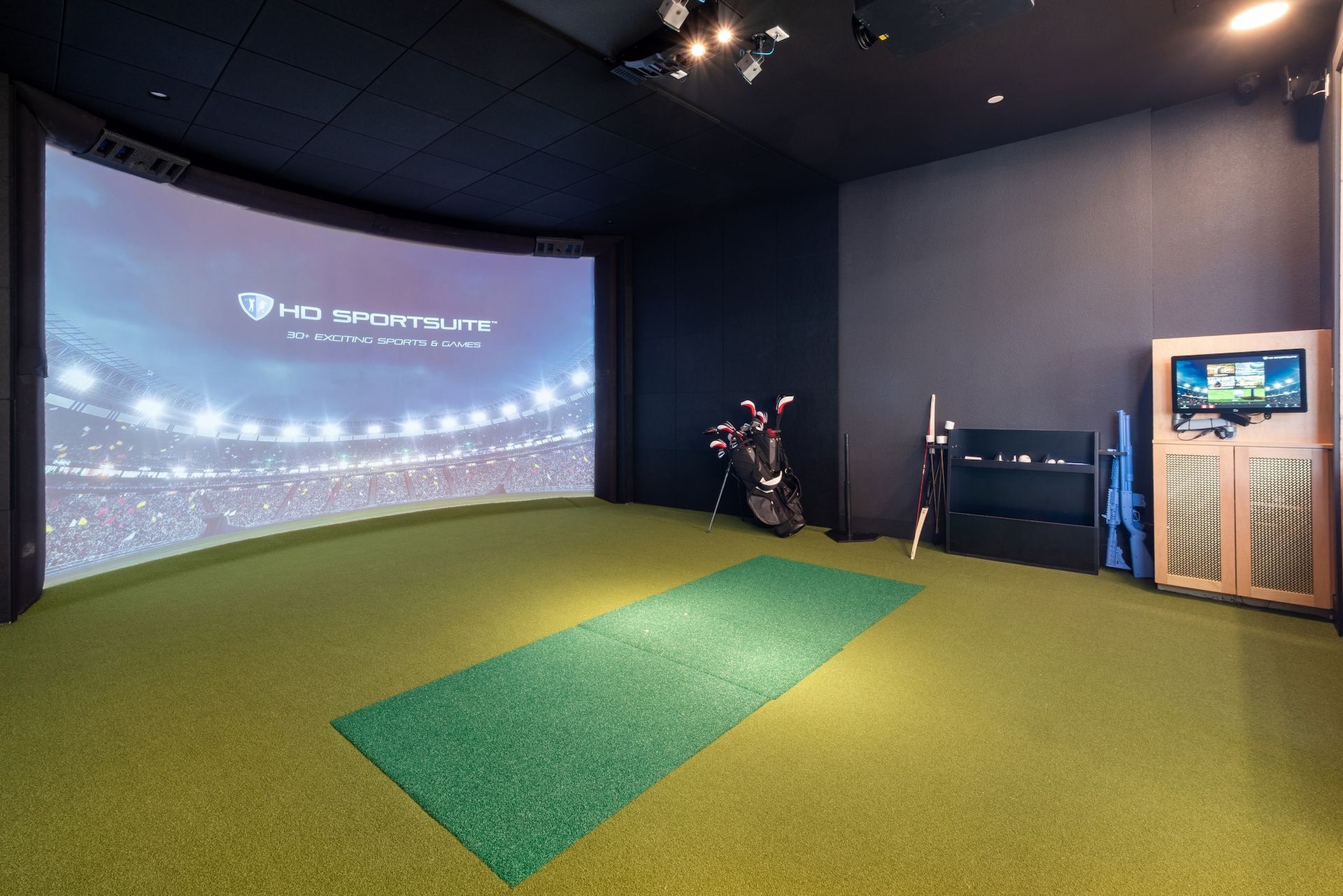 Golf simulator room with screen displaying a stadium, artificial turf, golf clubs, and a small entertainment console.