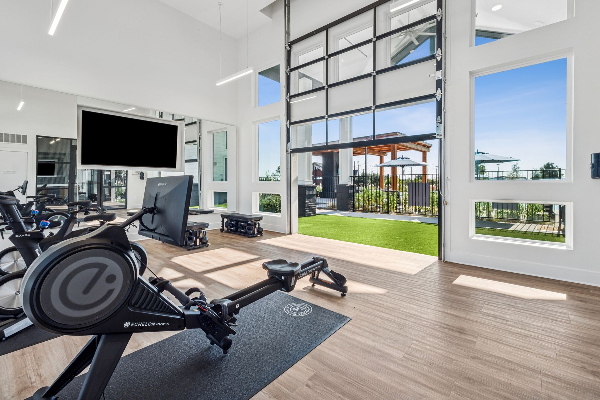Gym with row machine, large windows overlooking outdoor patio with green grass and pergola.