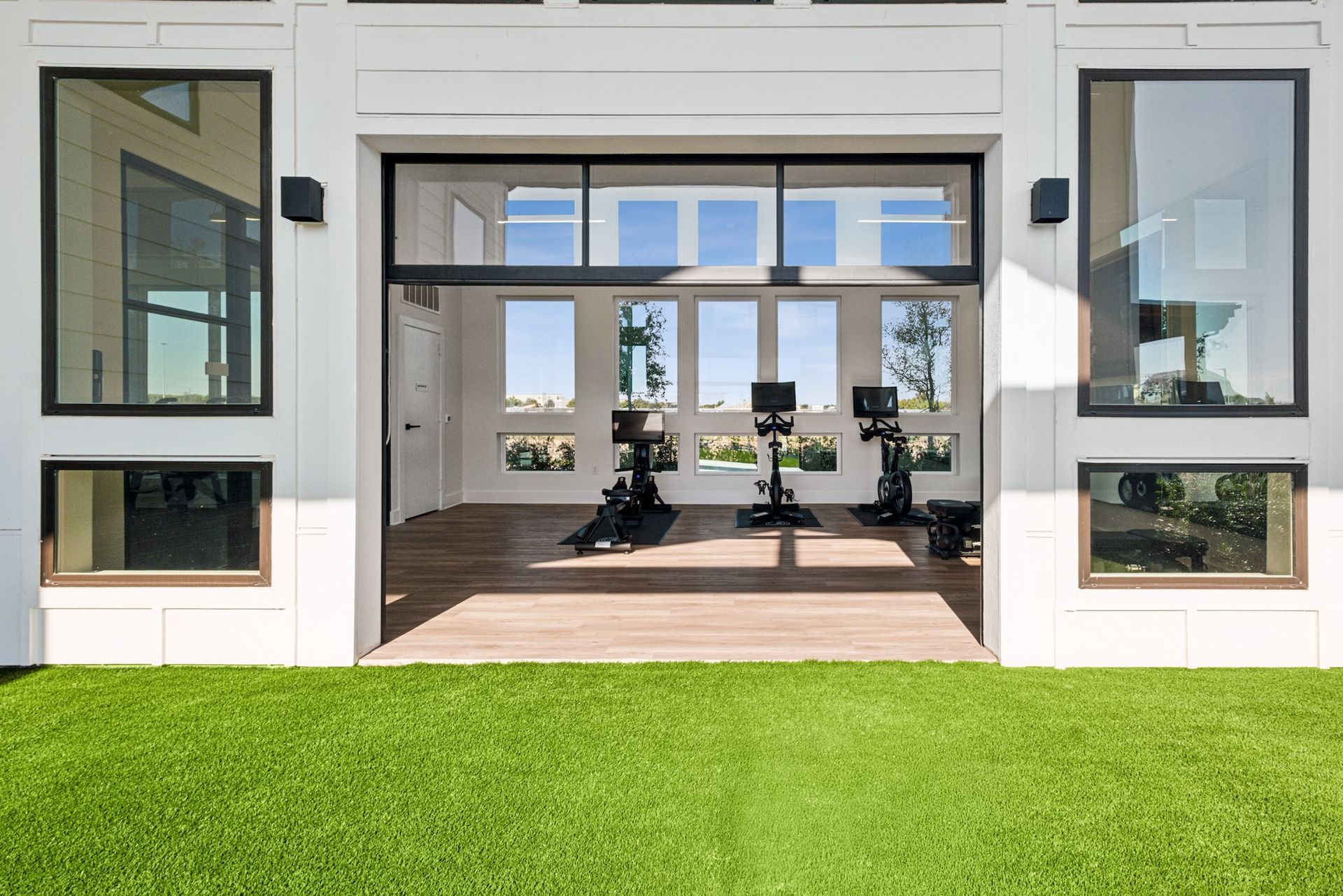 Exterior of a white home gym with exercise equipment visible through windows, set against a green lawn.